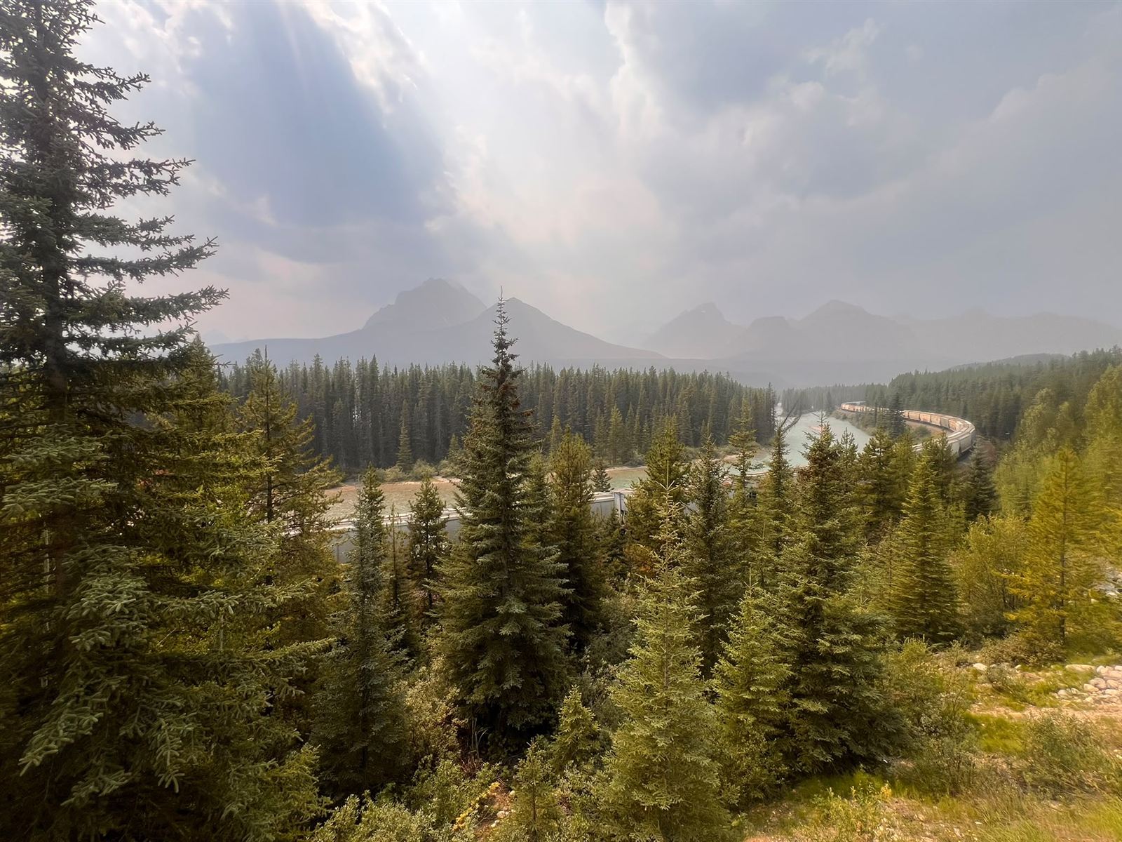 Wide view over evergreen forest to the Bow River and railway, with sunbeams breaking through cloud above the Bow Valley peaks