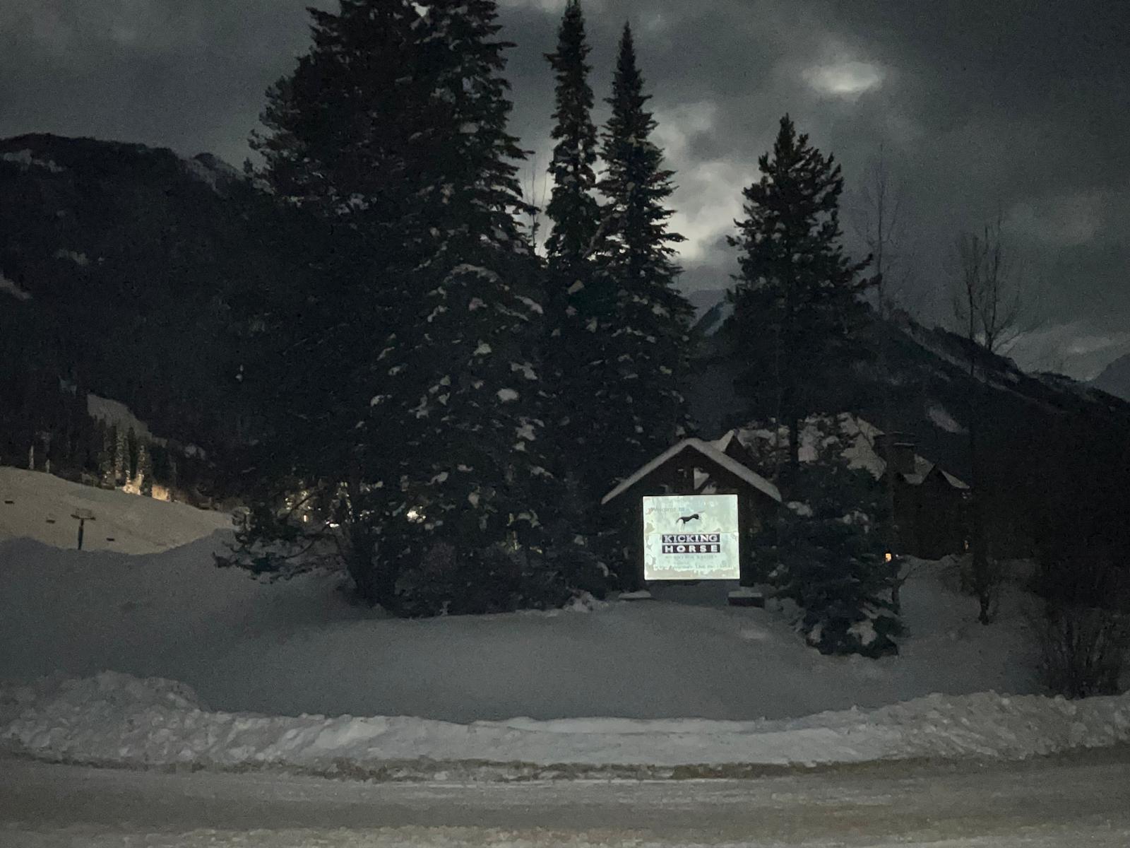 Illuminated Kicking Horse Mountain Resort welcome sign among snow-covered spruce trees at night